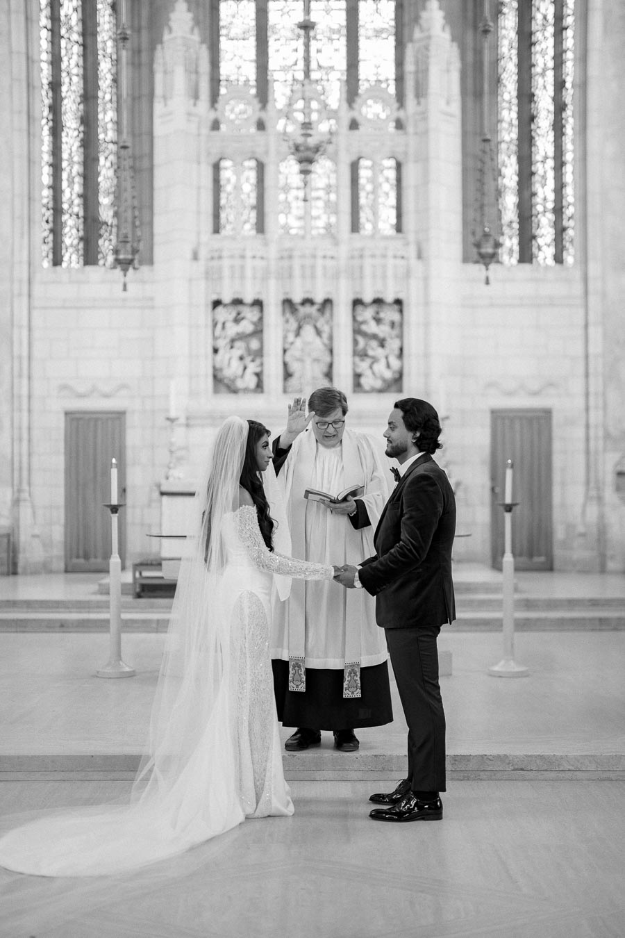 A bride and groom standing hand in hand during their wedding ceremony in a church, with an officiant reading from a book. The bride wears a long, flowing white gown and veil, while the groom is in a dark suit. The church's ornate altar and stained glass windows are visible in the background.