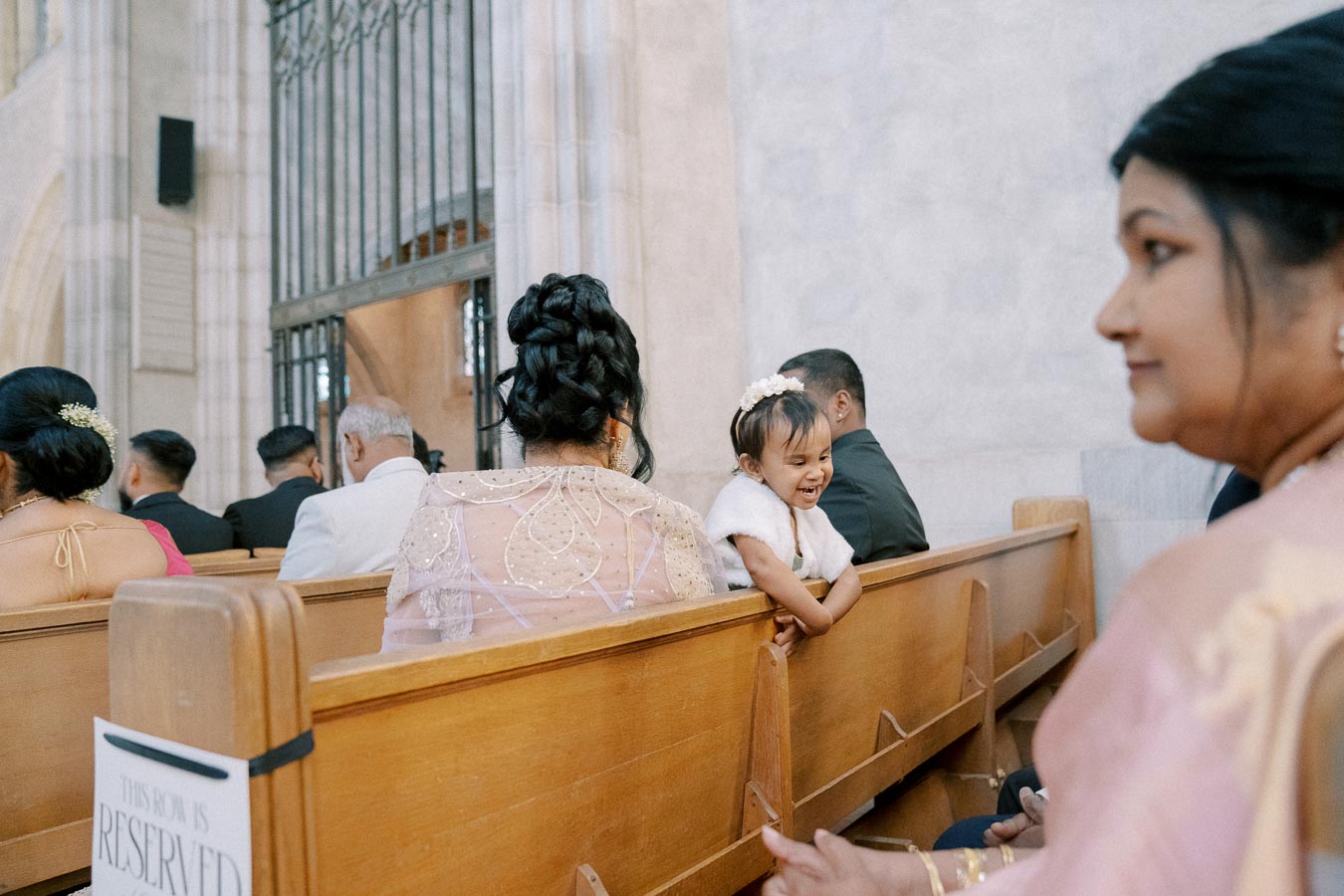 A joyful young girl in a white outfit smiles while leaning over a wooden pew during a formal ceremony in a grand church setting, surrounded by elegantly dressed guests.