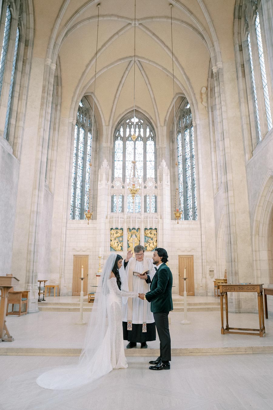 Elegant wedding ceremony in a grand cathedral with high vaulted ceilings and stunning stained glass windows, featuring a bride in a flowing white gown and veil holding hands with the groom in a dark suit, as a priest presides over the vows.