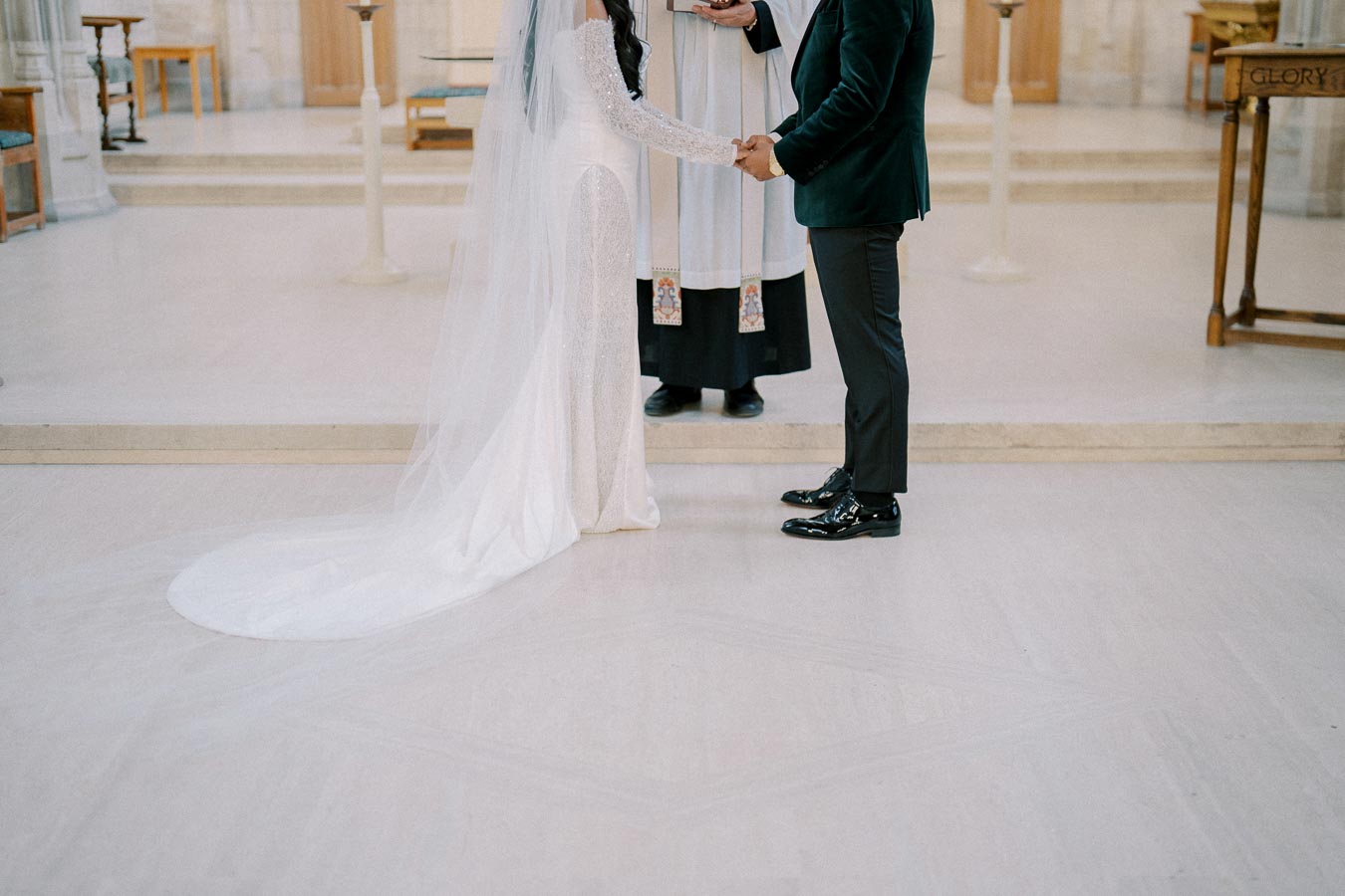 A bride and groom holding hands during their wedding ceremony in a church, with an officiant standing in the background. The bride is wearing a long white gown with a veil, and the groom is in a dark suit.