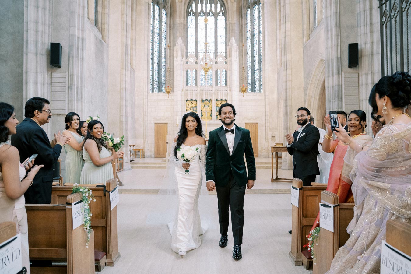 A joyful couple exits a beautifully adorned church aisle after their wedding ceremony, surrounded by cheering family and friends dressed in elegant attire, capturing a moment of celebration and happiness.