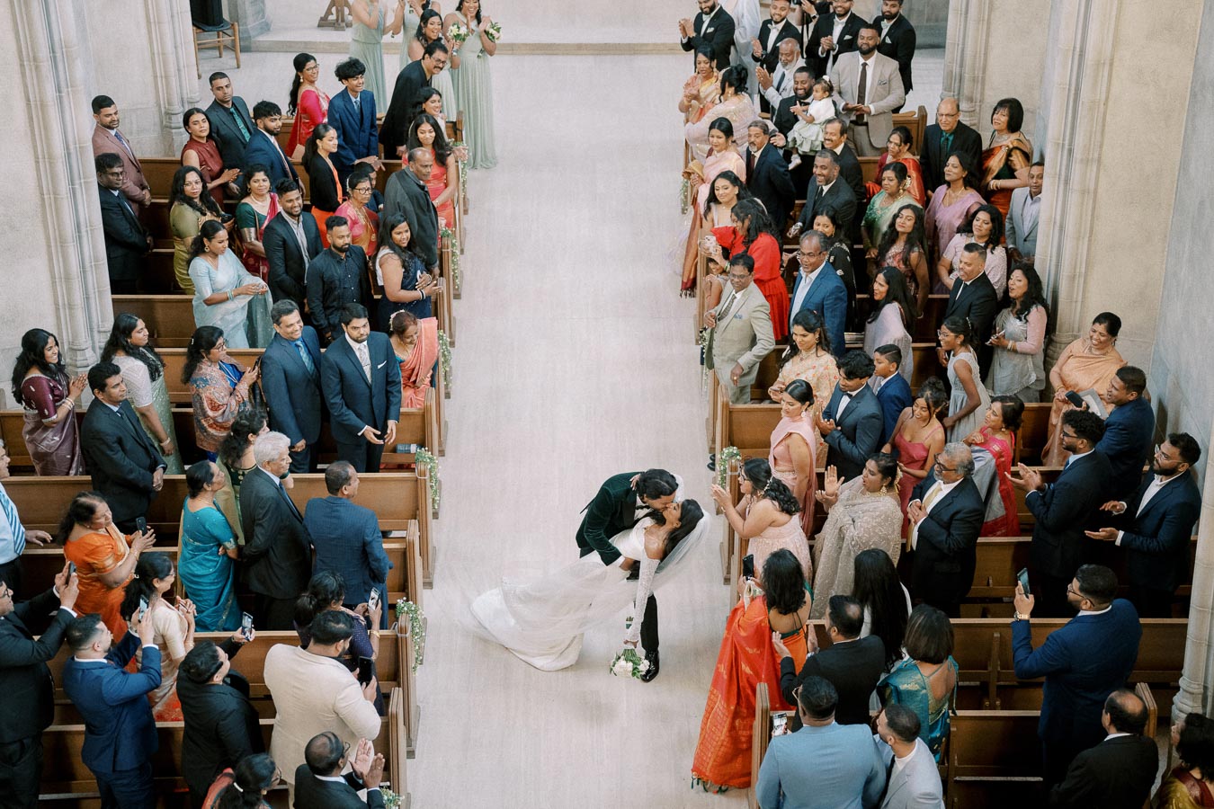 A couple shares a kiss at the end of a church aisle surrounded by applauding wedding guests during a multicultural ceremony.