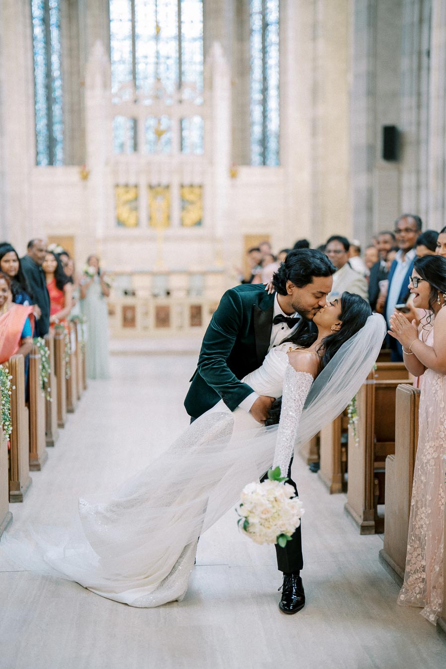 Bride and groom sharing a romantic kiss in a church aisle, with guests cheering in the background; the bride wears a flowing white gown and veil, and holds a bouquet of white flowers.