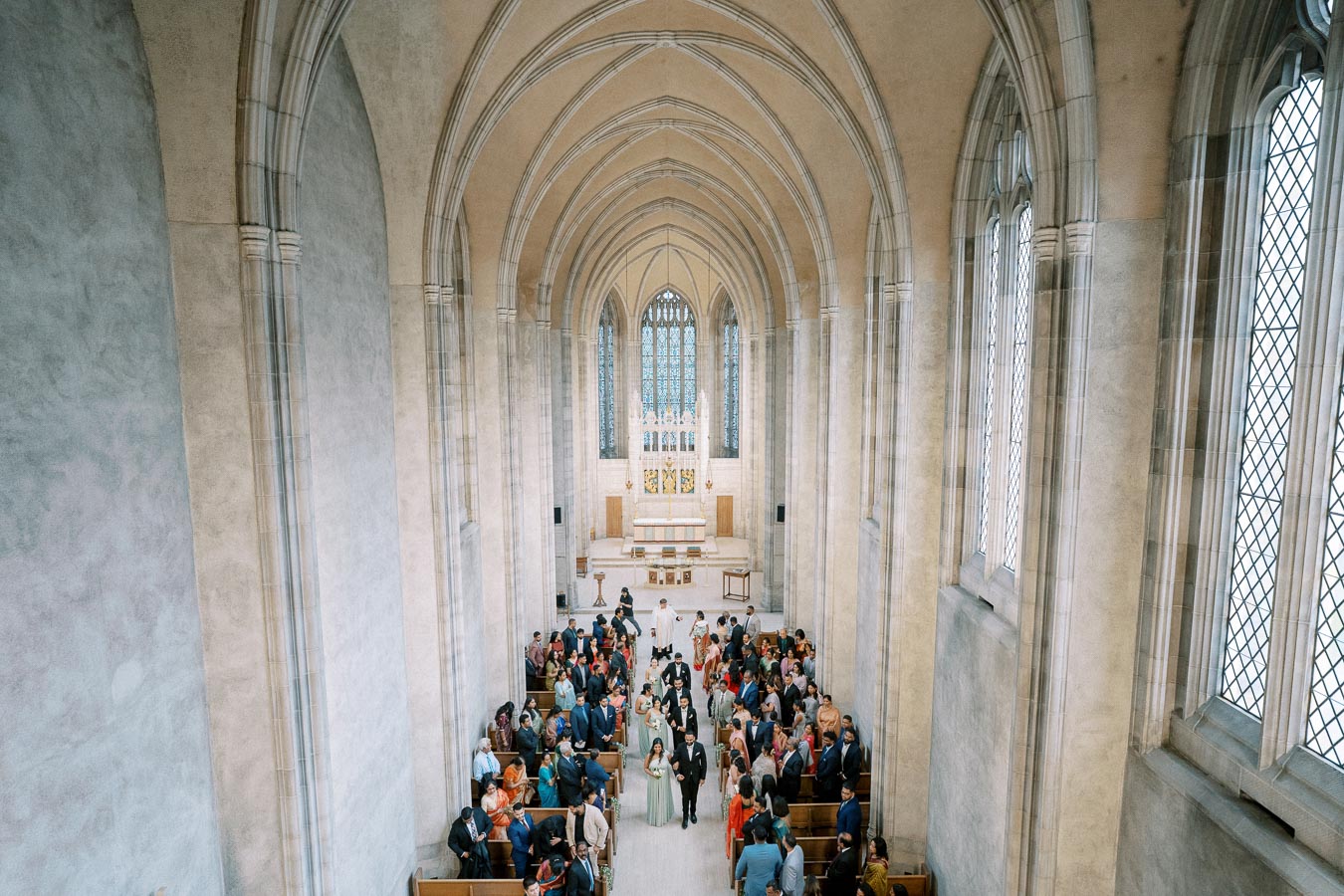 A wedding ceremony inside a stunning cathedral with tall arched ceilings, large stained glass windows, and a congregation of guests gathered in the pews.