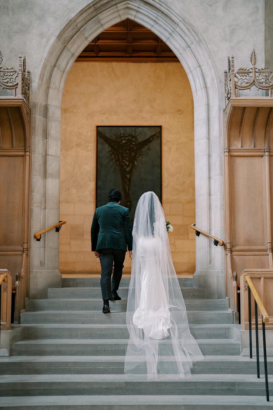A bride in a long, flowing white veil and gown walks up the stone steps of a grand historic building, followed by a groom in a dark suit. They are framed by an arched entrance and detailed architecture, with a large painting in the background.