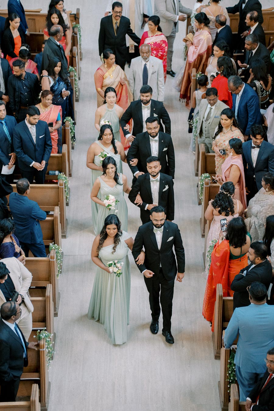 A wedding procession in a church with bridesmaids in light green dresses and groomsmen in black suits walking down the aisle, surrounded by seated guests dressed in colorful attire.