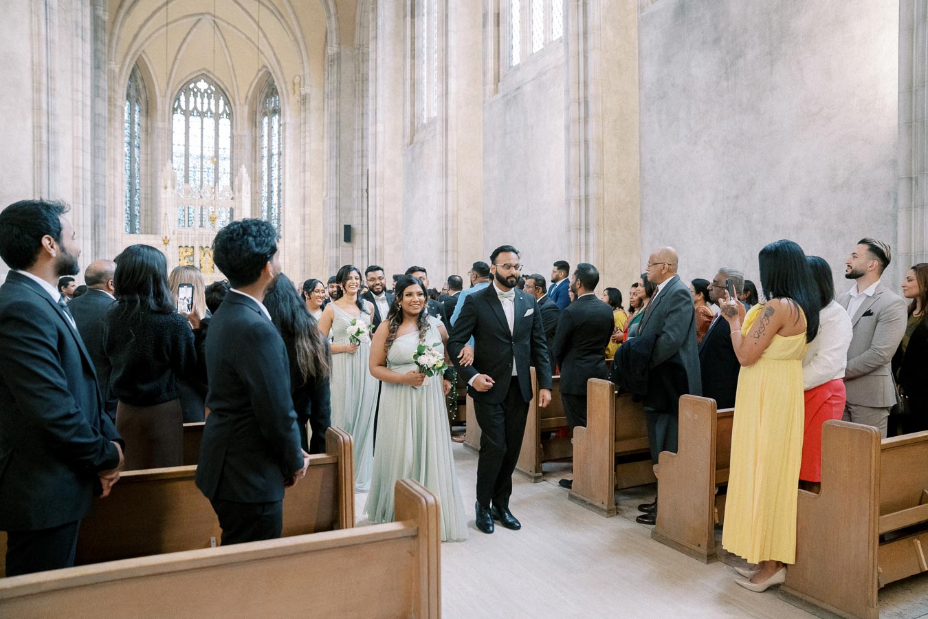 A wedding ceremony in a grand church with high ceilings, featuring a well-dressed man and woman walking down the aisle, surrounded by guests standing in pews. The woman is holding a bouquet and wearing a light green dress, while the guests capture the moment with their phones.