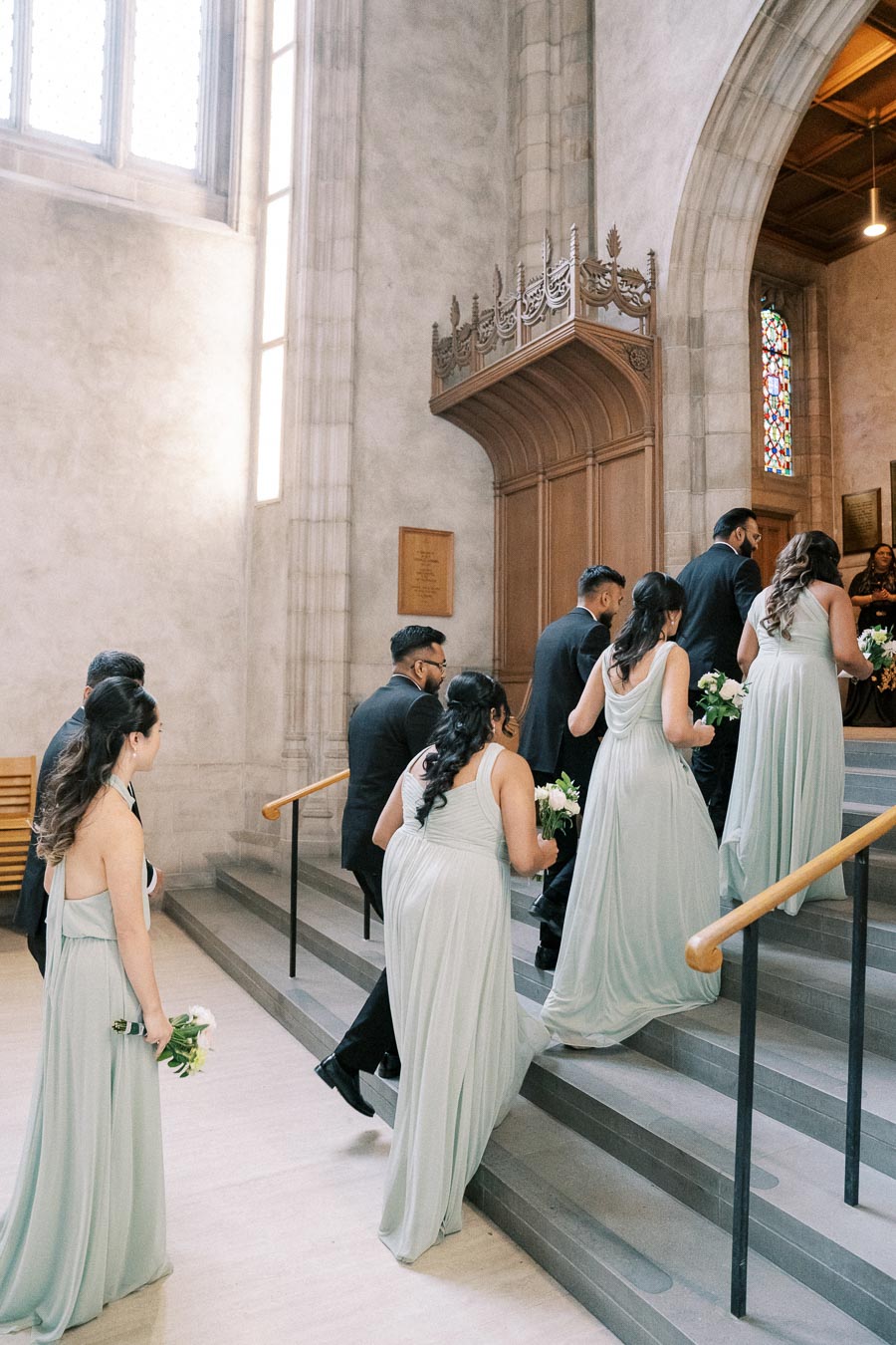 Bridal party in pale green dresses and black suits ascending stairs in a historic church for a wedding ceremony.