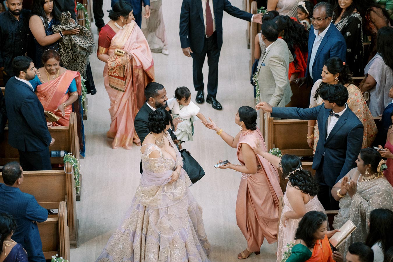 A joyful wedding ceremony scene with guests in colorful attire, including traditional saris and suits, surrounding the bride and groom holding a child. The indoor setting features wooden pews and decorative floral arrangements along the aisle.