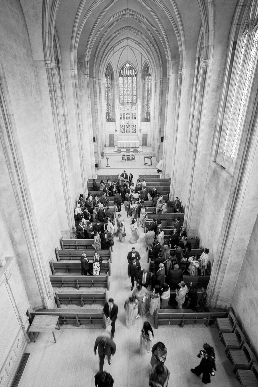 Black and white aerial view of a gathering in a large cathedral with high vaulted ceilings, showing people mingling in the pews and walking through the aisle during an event.
