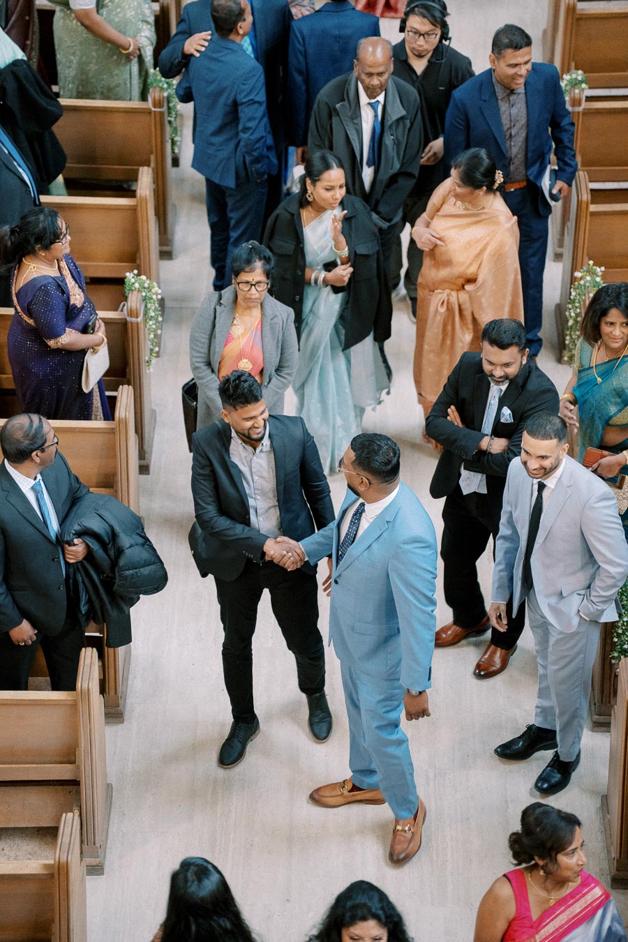 A diverse group of people dressed in formal attire gather and engage in conversation inside a church. Two men in suits prominently shake hands amidst the crowd, symbolizing connection and celebration.