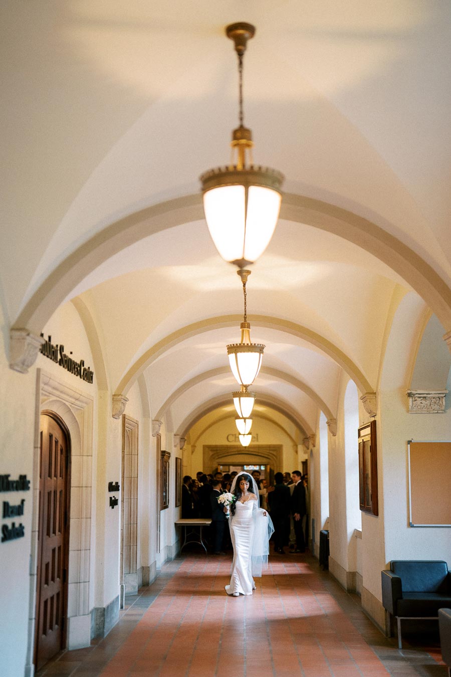 Bride walking down an elegant, arched hallway with hanging chandeliers, holding a bouquet, and wearing a white wedding dress.