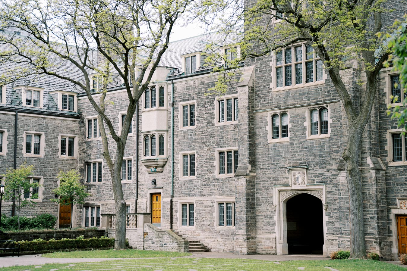 Historic stone university building with large windows and wooden doors, surrounded by greenery and trees, showcasing traditional collegiate architecture.