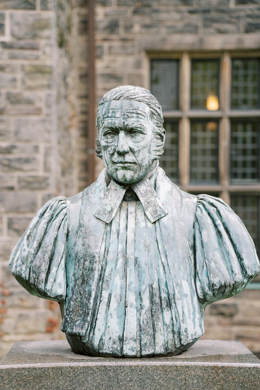 Bust of a historical figure in traditional attire, featured against a stone building background with windows.