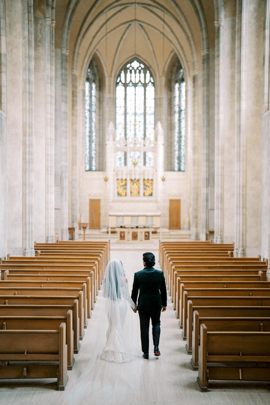 A bride and groom holding hands walk down the aisle of a grand cathedral, featuring high vaulted ceilings and ornate stained glass windows, capturing a serene and elegant wedding moment.