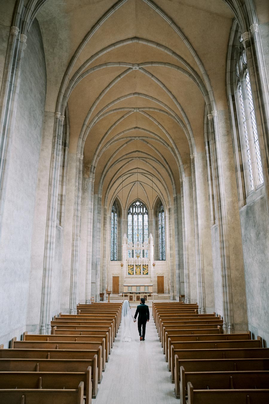 A bride and groom walk hand in hand down the aisle of a grand church with high vaulted ceilings and wooden pews, heading towards a beautifully ornate altar.