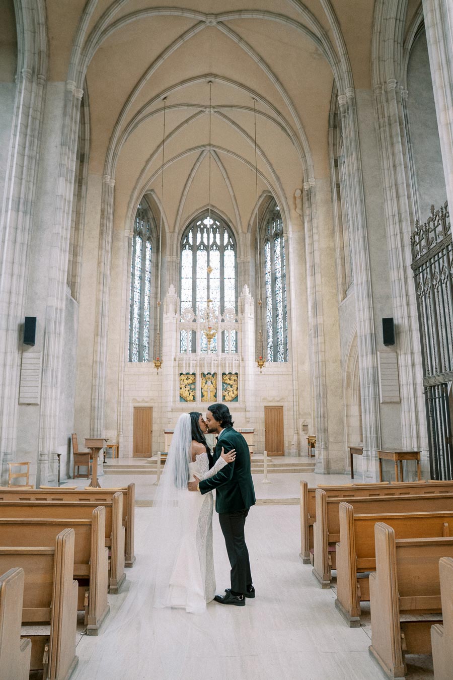 A bride and groom sharing a kiss in an elegant, high-vaulted church interior with gothic architecture, stained glass windows, and wooden pews.