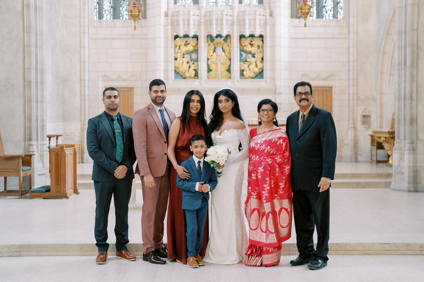 A diverse family group poses together in a church setting for a wedding. The bride in a white gown stands holding a bouquet, surrounded by family members dressed elegantly in suits and traditional attire. The intricate stained glass and detailed architecture provide a stunning backdrop.