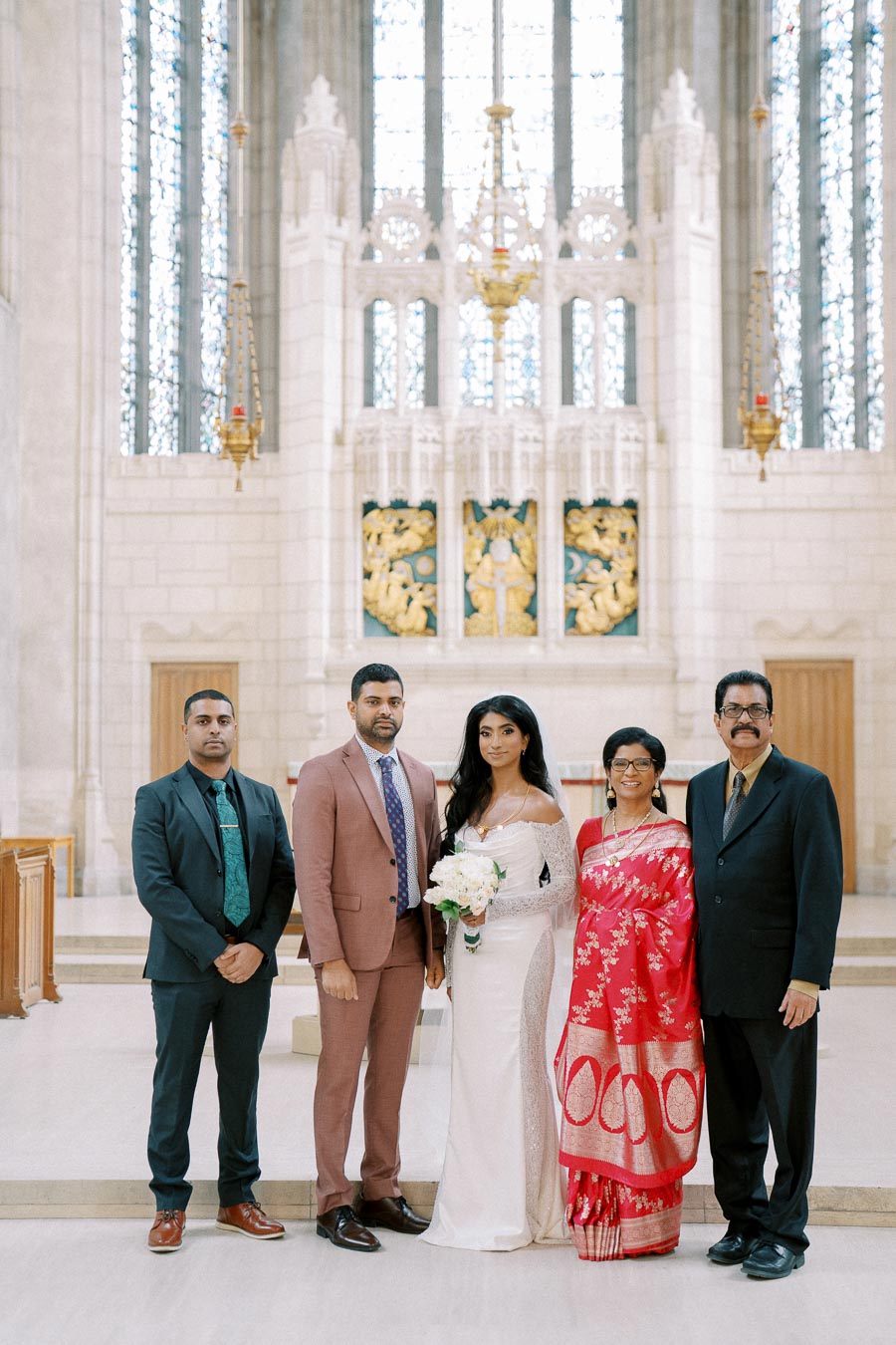Elegant wedding ceremony in a grand cathedral with beautifully decorated high ceilings, featuring a bride in a white dress holding a bouquet, flanked by family members in formal attire.