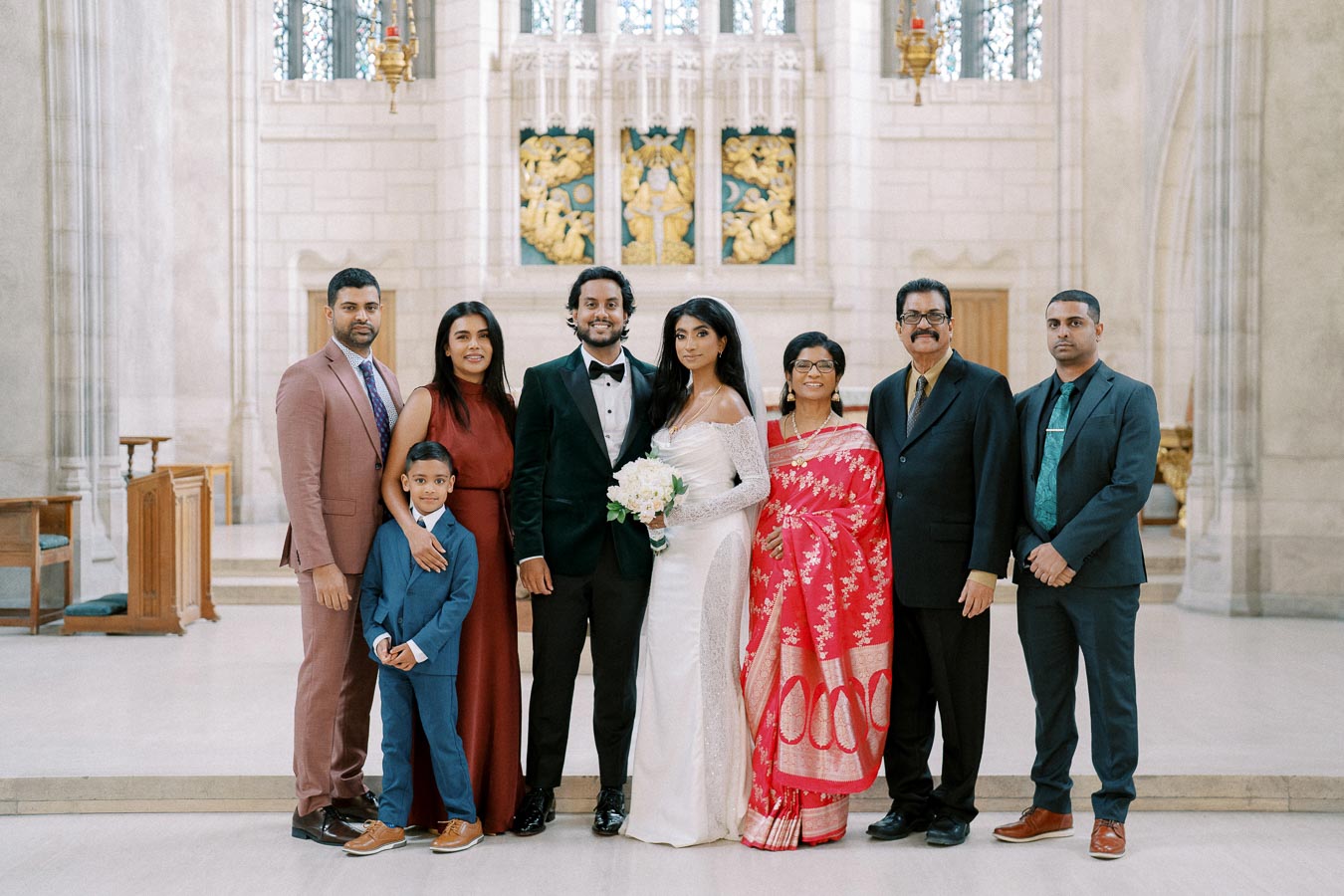 A group of people posing for a wedding photo inside a church, featuring the bride in a white dress and groom in a black suit, surrounded by family members dressed in formal attire with a mix of suits and a traditional red sari, standing in front of an ornate altar.