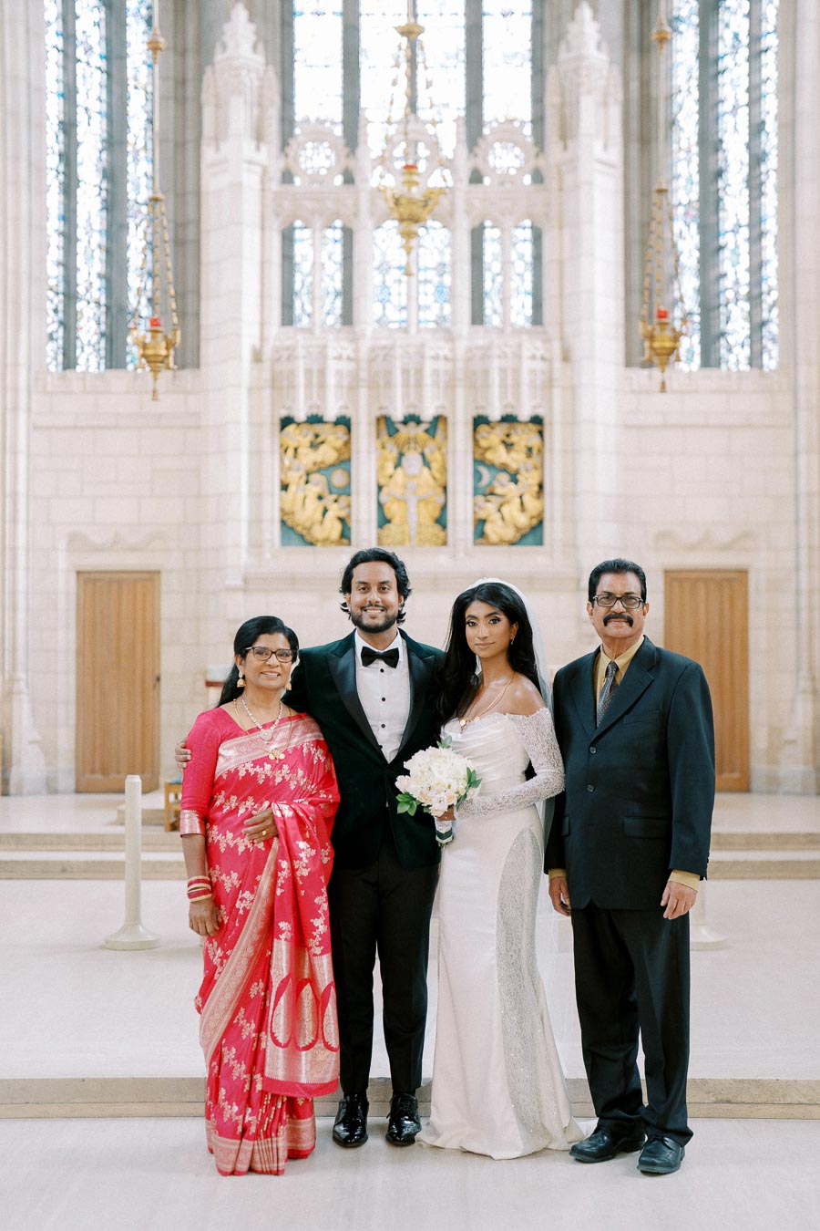 A newlywed couple poses with family in an elegant indoor setting, featuring a bride in a white wedding dress holding a bouquet and a groom in a black tuxedo. The backdrop showcases intricate architectural details and stained glass windows. One family member is wearing a vibrant red saree, and another is in a dark suit.