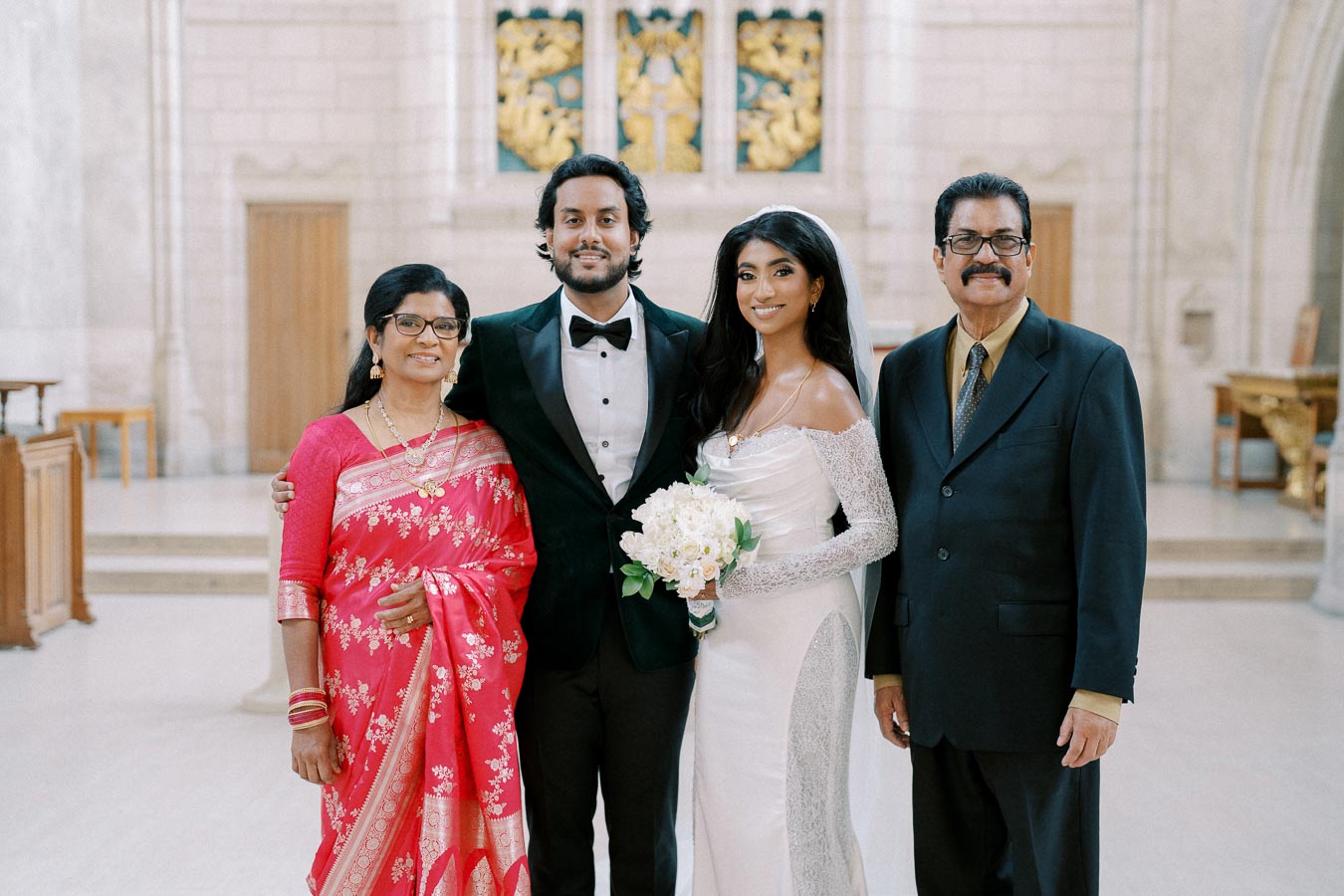 A family posing together indoors during a wedding ceremony. The groom in a tuxedo and the bride in a white wedding dress stand in the center, holding a bouquet. The bride’s parents are beside them, with the mother wearing a vibrant pink saree and the father in a dark suit. The background features ornate architectural details.