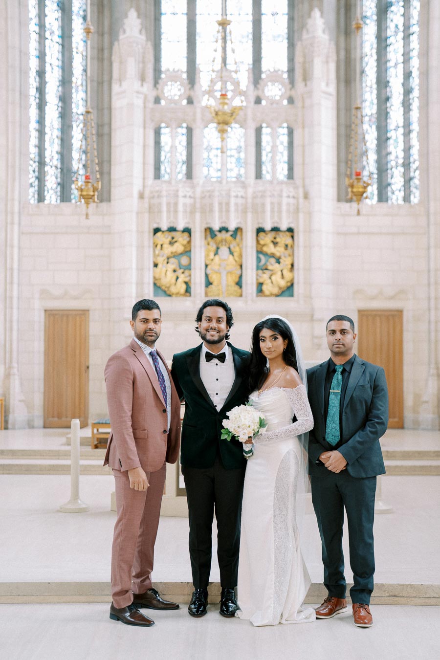 Elegant wedding ceremony in a grand cathedral with a bride in a beautiful white gown and groom in a tuxedo, flanked by two groomsmen in stylish suits, posing together before a dramatic altar backdrop.