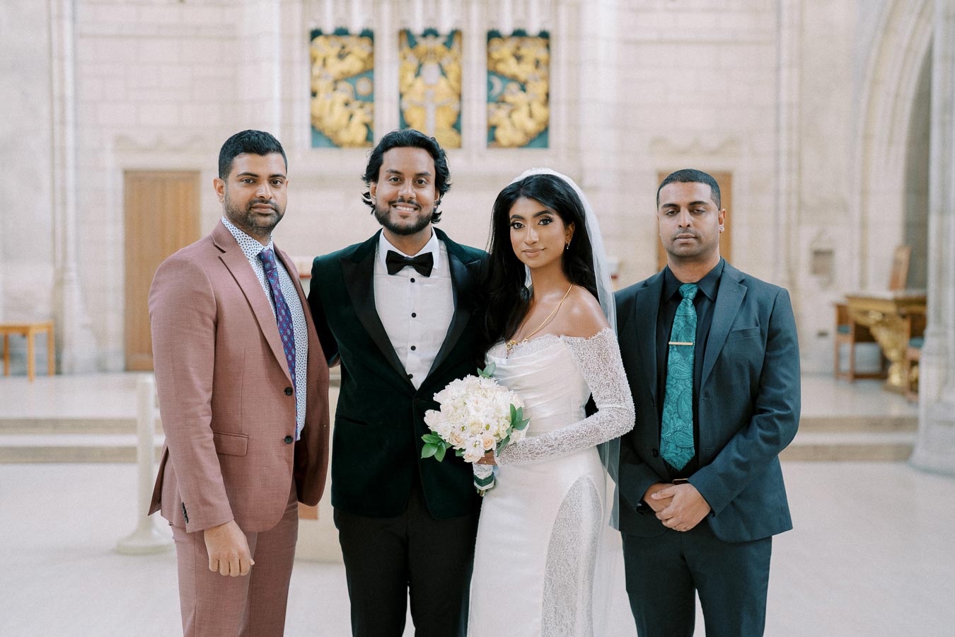 A bride and groom pose with two groomsmen at a wedding inside a grand, elegantly decorated venue. The bride is wearing a lace wedding dress and holding a bouquet, while the groom and groomsmen are dressed in formal suits.