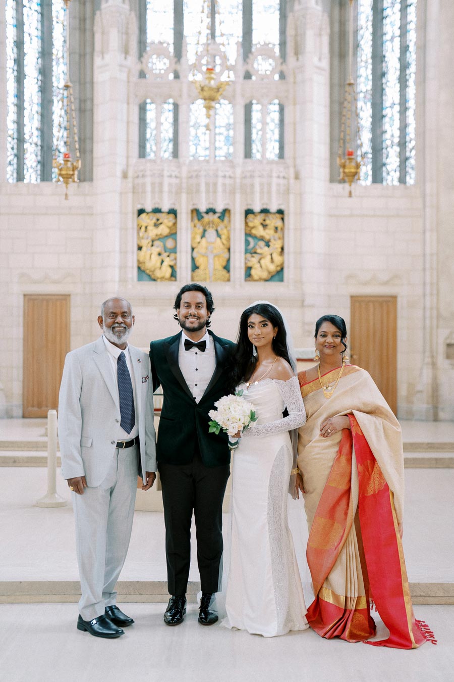 A newlywed couple posing with their parents in an elegant church setting, featuring the bride in a white gown holding a bouquet, the groom in a tuxedo, with the parents in formal attire including a sari.