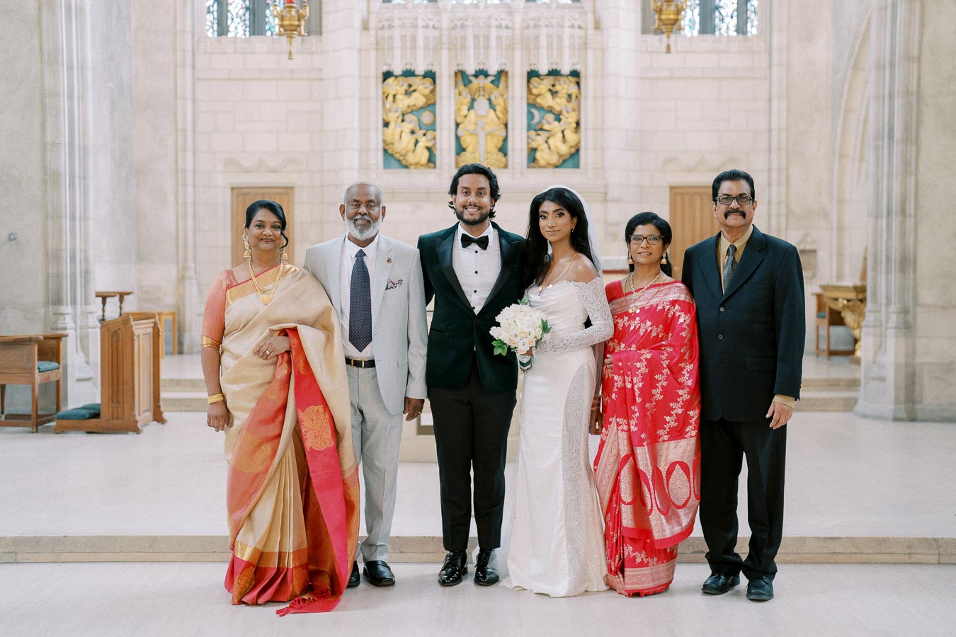 A wedding photo featuring a newlywed couple with their family members in traditional and formal attire, standing in a beautifully decorated church setting. The bride is in a white gown holding a bouquet, while the groom wears a classic tuxedo. Family members are dressed in vibrant saris and suits.