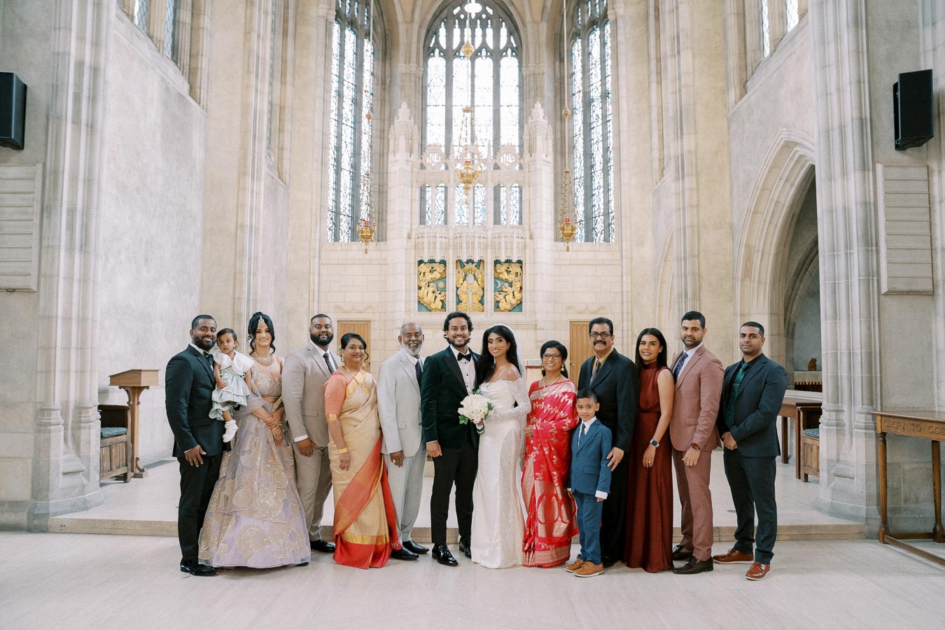 A diverse group of well-dressed people posing for a formal group photo in an ornate church interior with high ceilings and large stained glass windows. The central couple stands out, with the bride holding a bouquet.
