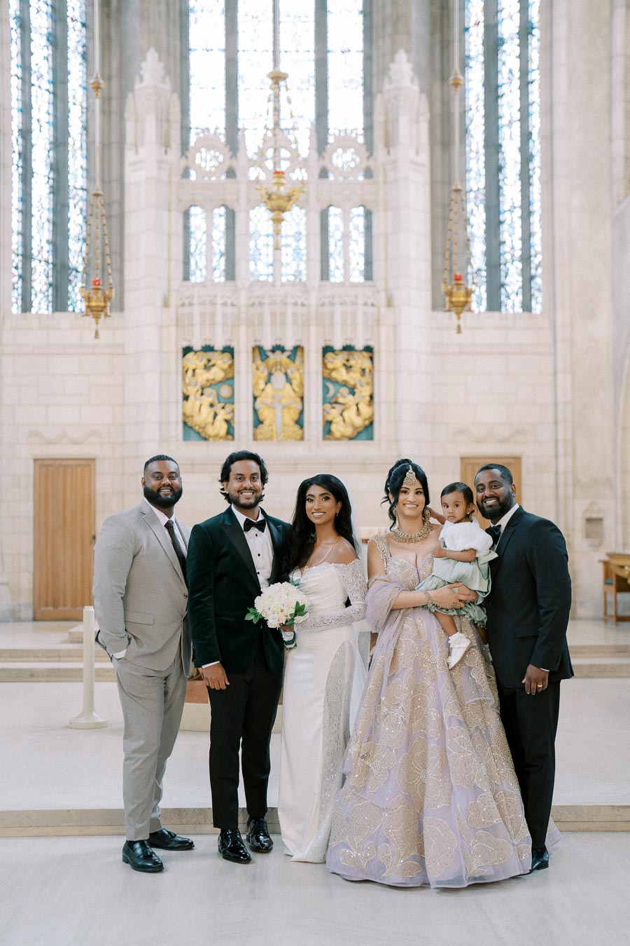 A group of elegantly dressed individuals posing in a grand church setting, featuring a bride in a white gown, a groom in a black tuxedo, and guests in formal attire. The backdrop includes ornate architecture and stained glass windows, adding to the sophisticated atmosphere of the event.