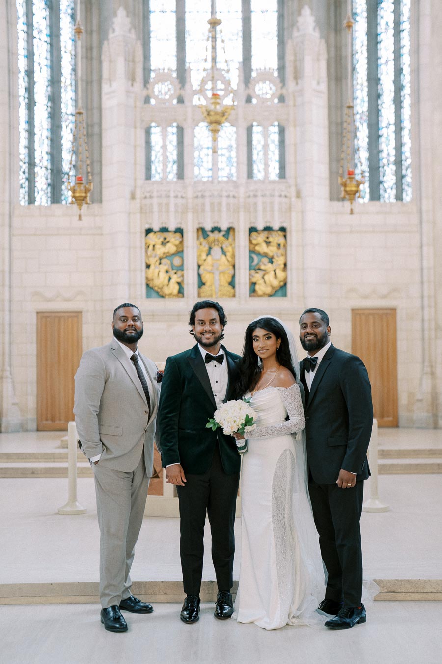 A bride in an elegant white gown and groom in a green velvet tuxedo pose with two groomsmen in a beautiful cathedral setting with intricate gold and blue details on the walls.