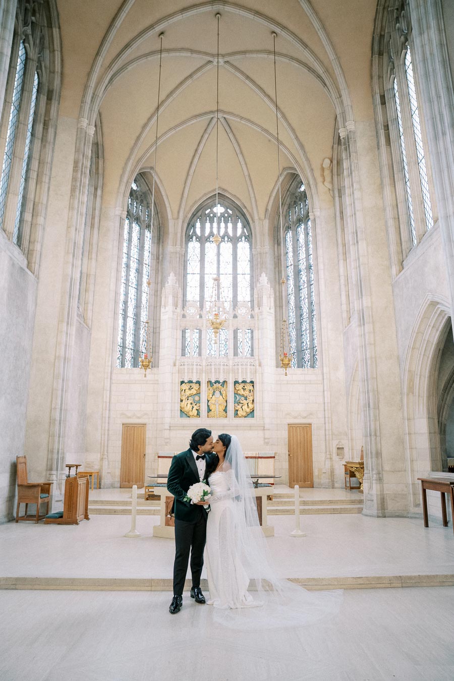 A newlywed couple kisses inside a grand cathedral with high vaulted ceilings and stained glass windows. The bride is wearing a traditional white wedding gown and veil, while the groom is in a classic black tuxedo. They are holding a bouquet of white flowers, as soft, natural light illuminates the elegant architectural details of the church.