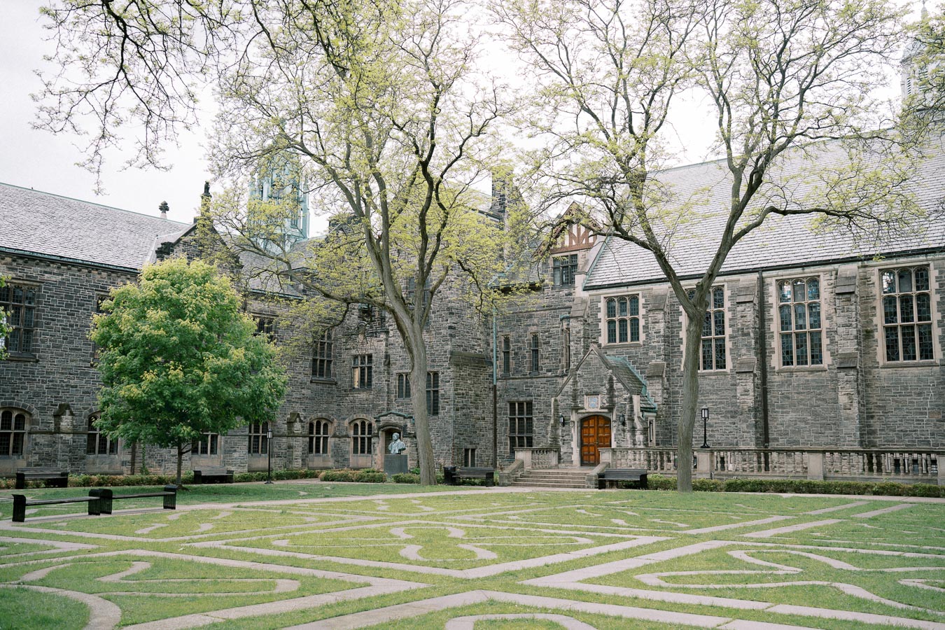 Historic stone university building with courtyard and geometric garden design, framed by tall trees.