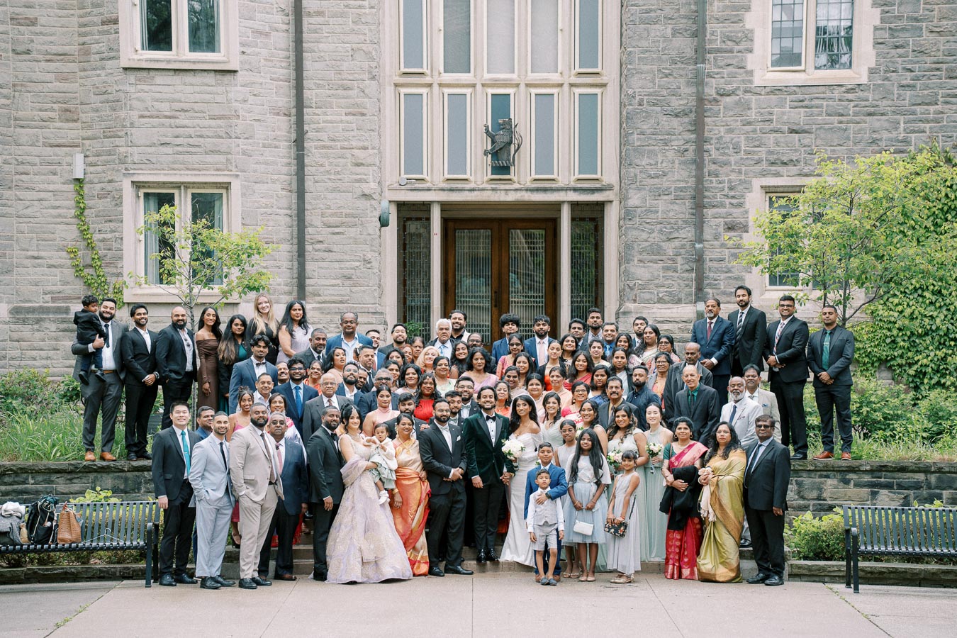 A large group of people in formal attire gathered for a wedding photo in front of a historic stone building, with greenery and trees surrounding the scene.