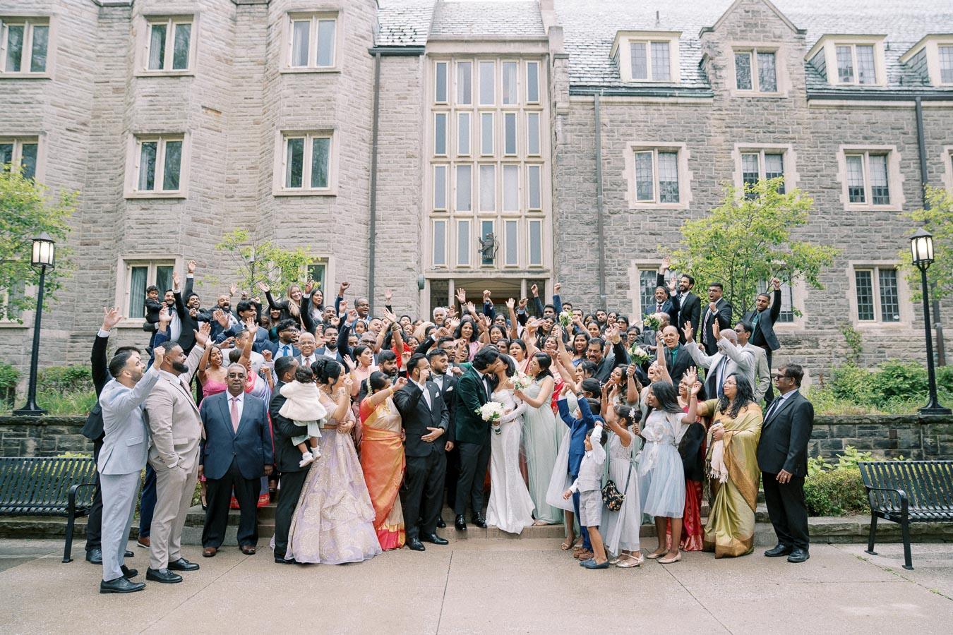 Joyful wedding celebration with a large group of guests posing in front of a historic stone building, featuring the bride and groom kissing in the center while friends and family cheer around them, dressed in colorful formal attire.