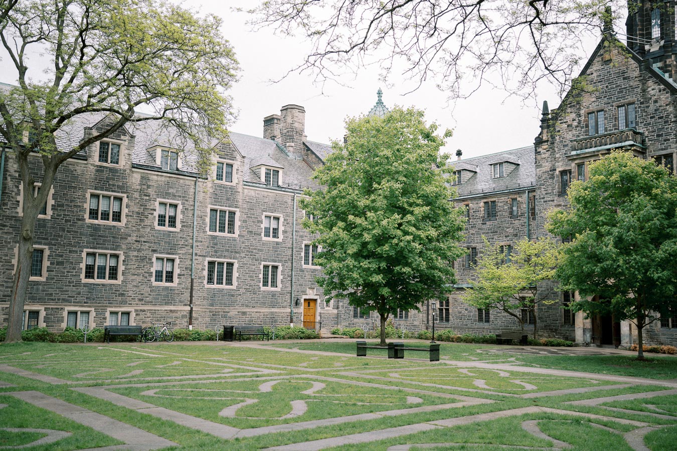 University campus courtyard featuring a historic stone building with multiple windows, surrounded by lush green trees and well-maintained grass pathways.