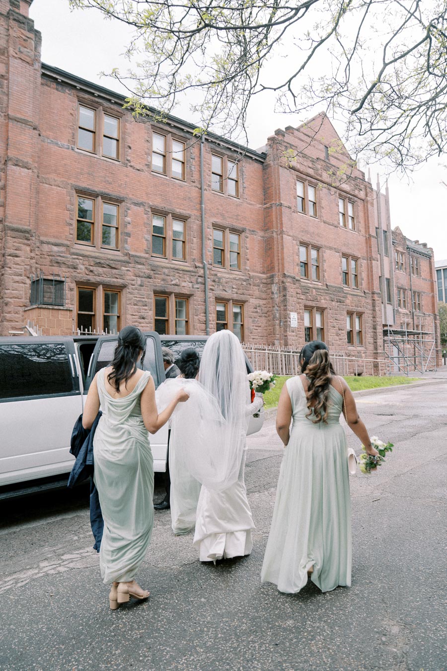 Bridal party walking towards a white limousine parked by a historic red brick building, capturing a moment of a wedding day.
