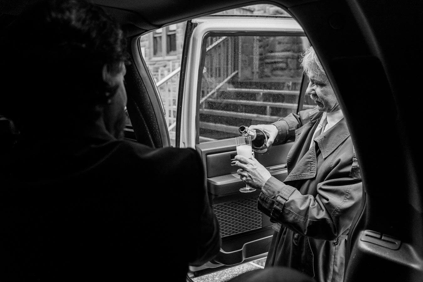 Black and white image of a person in a trench coat pouring champagne into a flute through a car door, with a blurred background of steps outside.