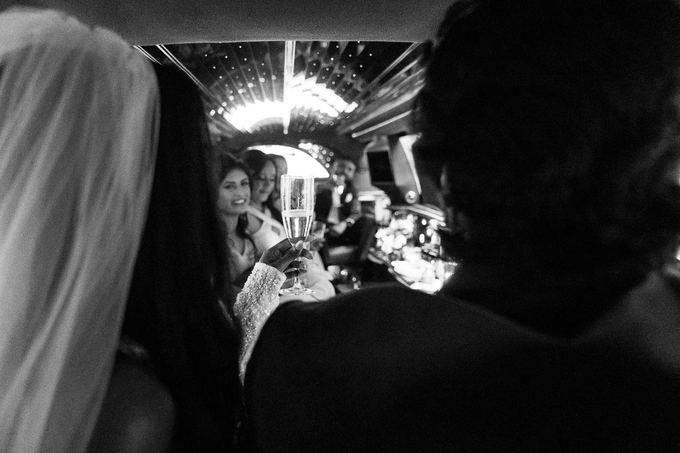 A black-and-white image of people celebrating inside a luxury limousine, with a focus on a hand raising a champagne glass. The ambiance is elegant, enhanced by illuminated ceiling lights, suggesting a festive occasion such as a wedding or party.