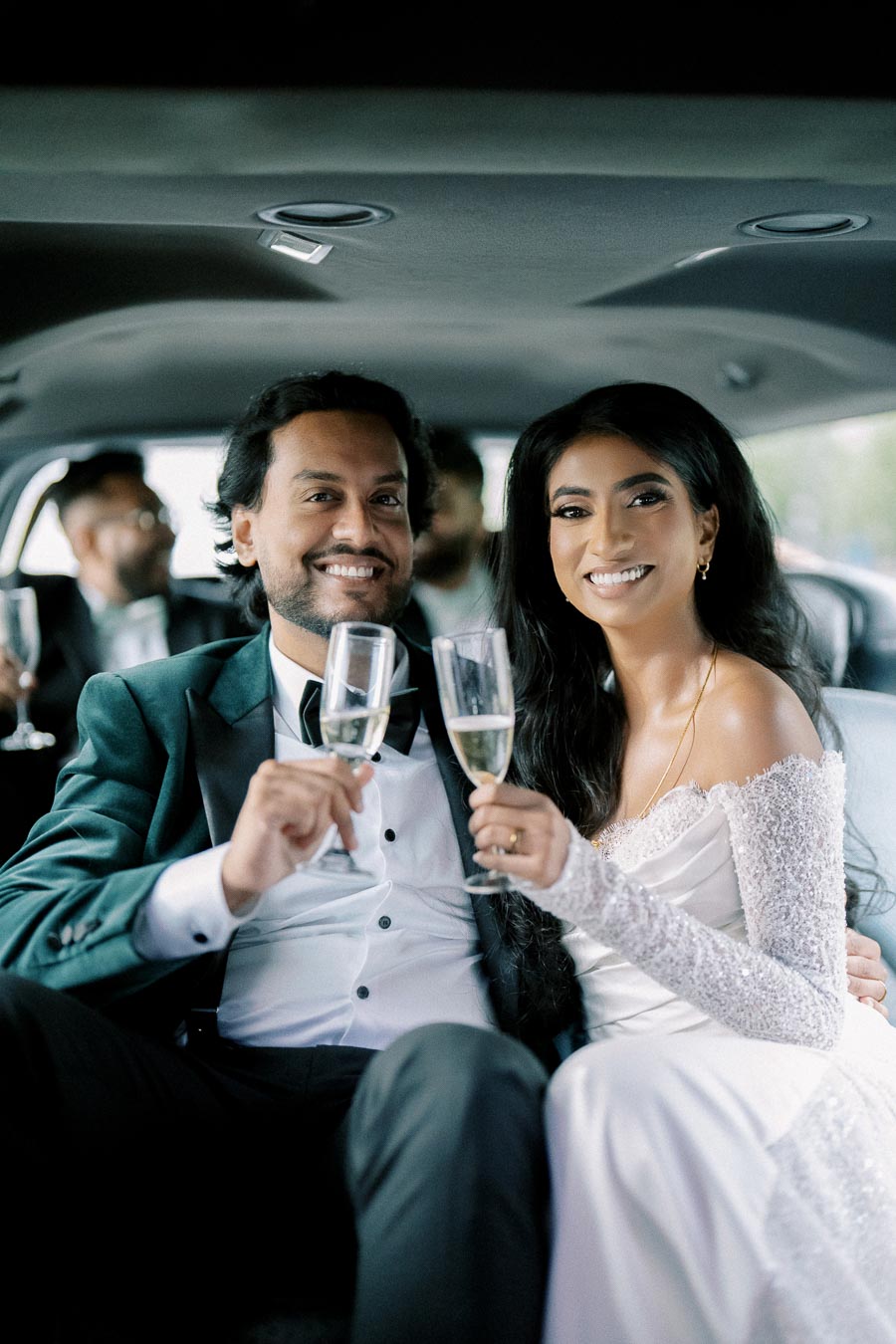 Joyful couple celebrating in a limousine, elegantly dressed and smiling, holding champagne glasses on their wedding day.