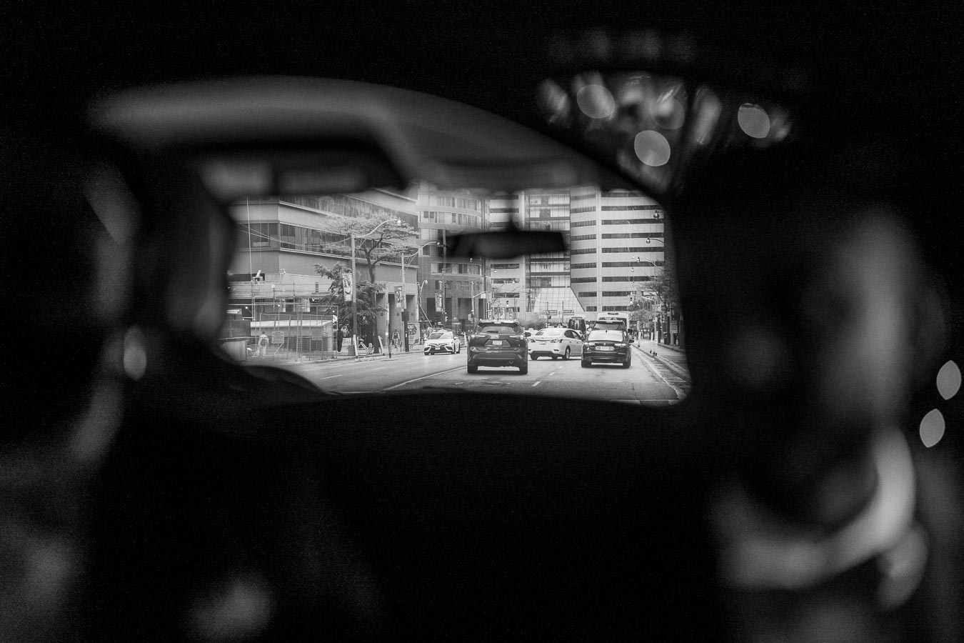 Black and white image of a busy urban street taken from inside a vehicle, with modern buildings and several cars visible on the road, framed by the car's rear-view mirror, creating a cinematic perspective.