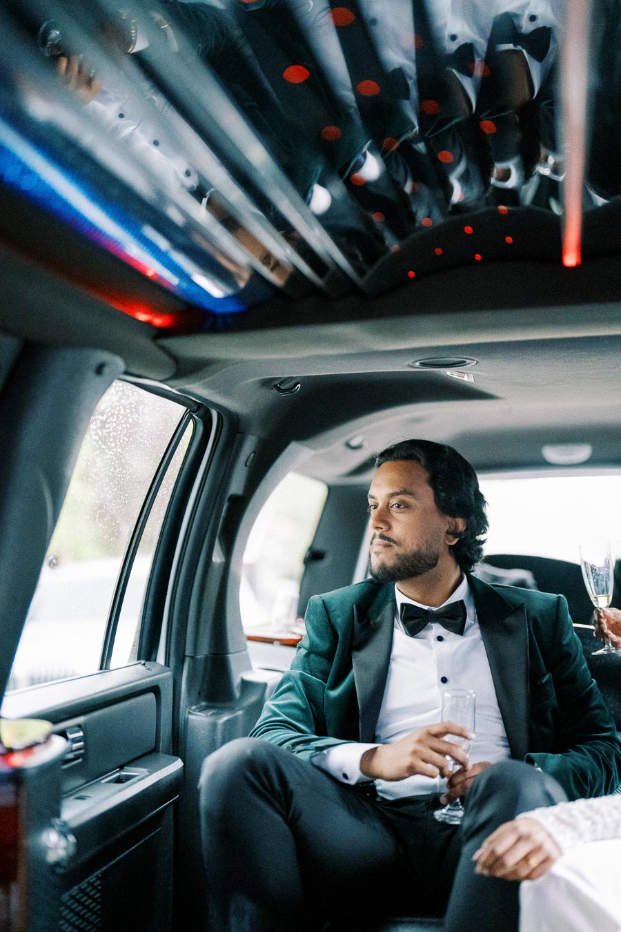 A well-dressed man in a tuxedo sits in the luxurious interior of a limousine, holding a glass, with ambient lighting and reflections creating a sophisticated atmosphere.