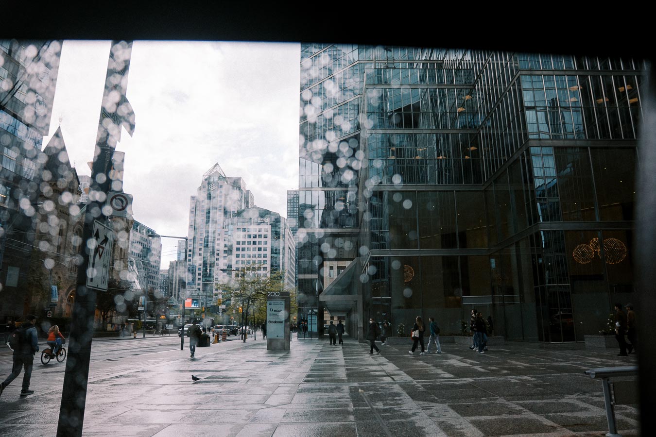 Rainy cityscape view through wet window glass showing pedestrians and cyclists on a busy urban street with modern glass buildings in the background.