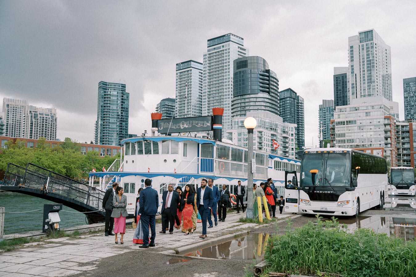 A group of people dressed in formal attire gather at a harbor near a docked tour boat and parked buses, set against the backdrop of modern high-rise buildings under an overcast sky.