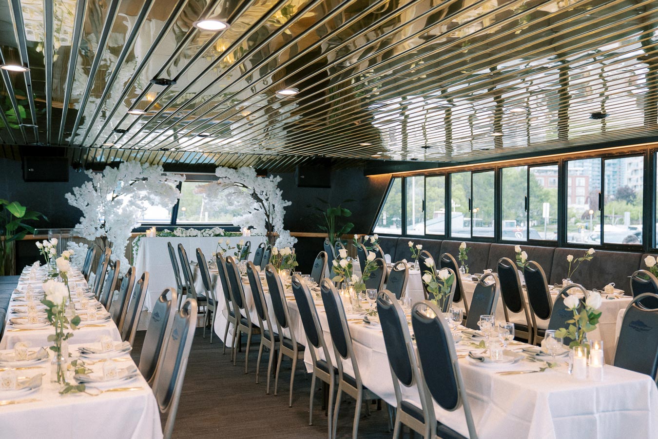 Elegant event setup in a tastefully decorated banquet hall with long tables dressed in white tablecloths, adorned with white floral centerpieces, and surrounded by navy blue chairs under a modern metallic ceiling, offering a luxurious dining experience.