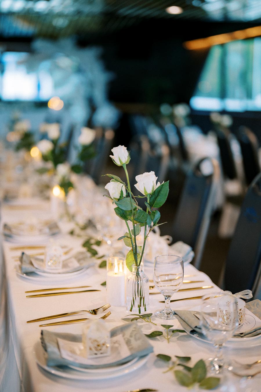 Elegant wedding table setting with white roses in glass vases, gold utensils, and candlelit ambiance.