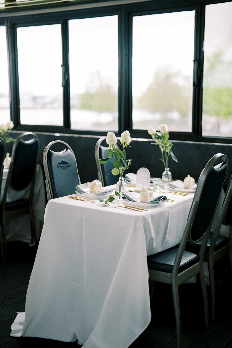 Elegant banquet table with white tablecloth, adorned with white roses and gold cutlery, set near large windows with a scenic view.