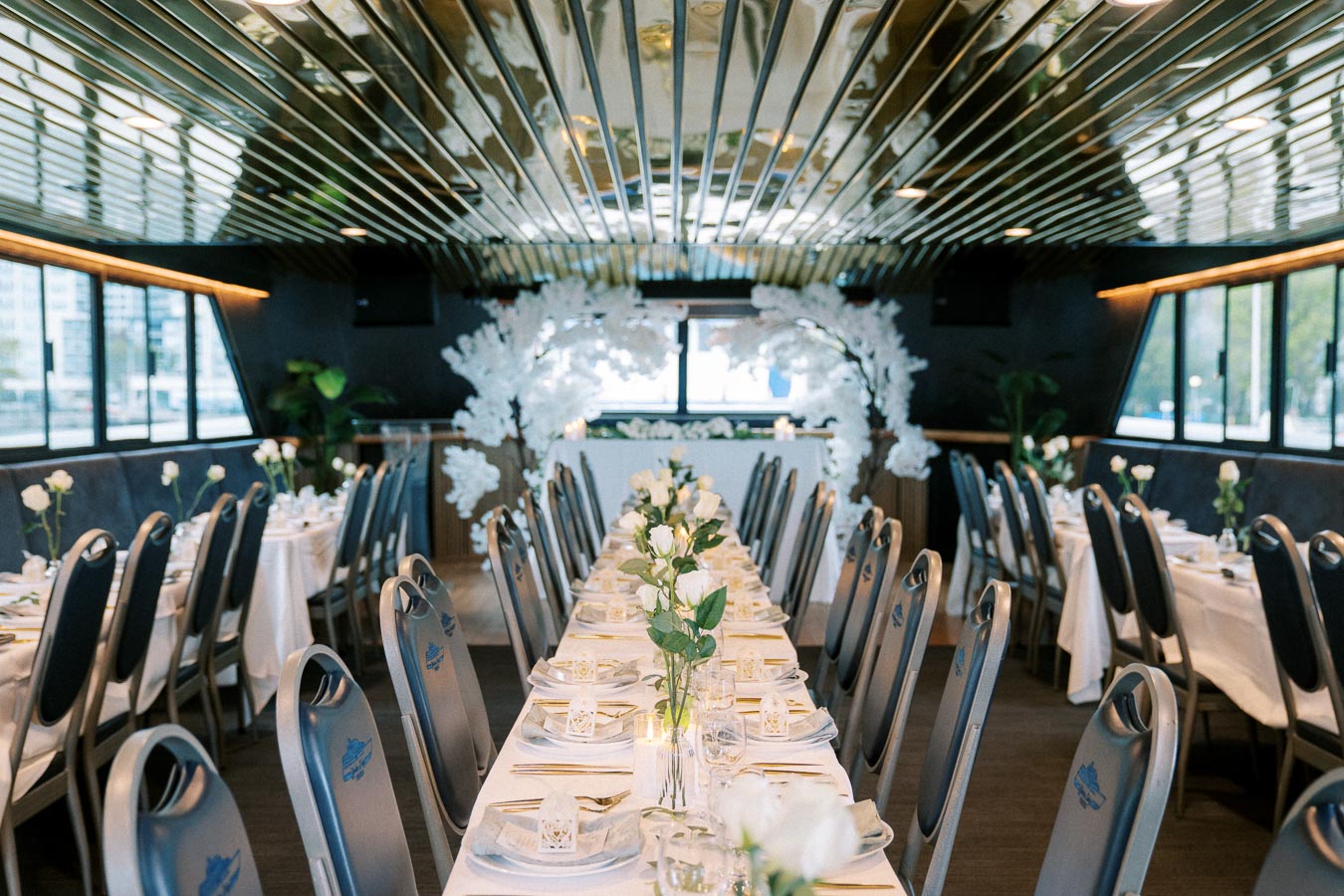 Elegant dining setup aboard a modern event cruise ship with rows of tables decorated with white roses and fine dining settings, featuring a stylish floral arch centerpiece and panoramic windows.